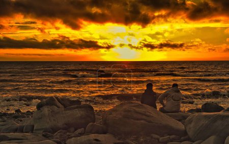 Téléchargez les photos : Deux personnes assises sur les rochers de la mer et regardant le coucher de soleil orange dramatique - en image libre de droit