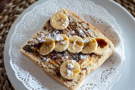 Foto de Un primer plano horizontal de plátanos recién cortados, chocolate y azúcar en polvo sobre pan tostado en un plato blanco - Imagen libre de derechos
