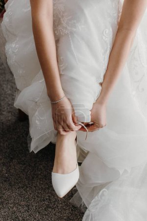 Foto de Una novia en un vestido de novia elegante blanco que se pone sus zapatos para el día de la boda - Imagen libre de derechos