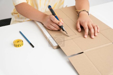 Téléchargez les photos : Une fille découpe du carton avec un couteau sur une épaisse surface blanche sur un fond blanc - en image libre de droit