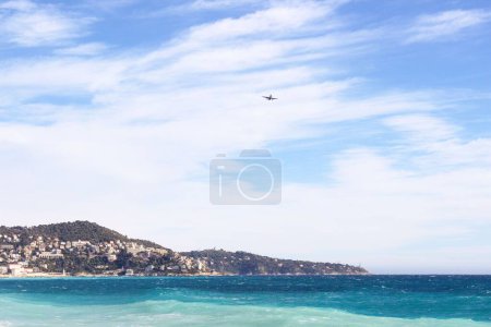Téléchargez les photos : Une vue de la mer de la ville à flanc de colline sur la côte avec un avion volant dans un ciel nuageux bleu - en image libre de droit