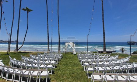 Foto de Una vista panorámica de sillas blancas en fila sobre hierba verde listo para una ceremonia de boda en la playa - Imagen libre de derechos