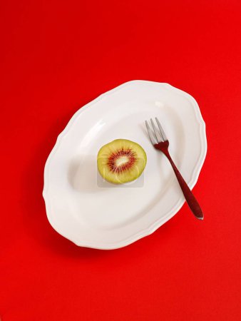 Photo for A vertical shot of a half of kiwi fruit in a white plate with a fork on red background - Royalty Free Image