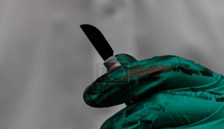 Téléchargez les photos : Une main dans un gant en plastique vert tenant un scalpel couvert de sang - en image libre de droit