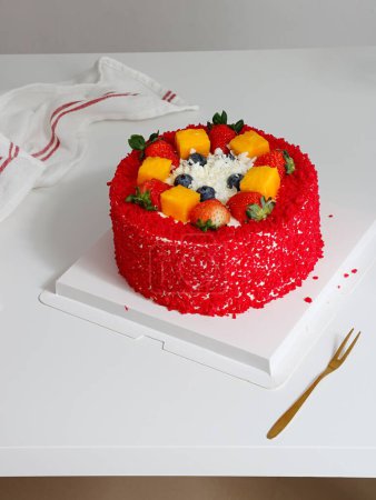 Téléchargez les photos : Un gâteau rond rouge décoré de bleuets, fraises et mangue avec un verre d'eau à l'arrière - en image libre de droit