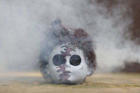 Téléchargez les photos : Une tête de poupée effrayante sur le gorund, avec des boutons à la place des yeux, entourée de fumée - en image libre de droit