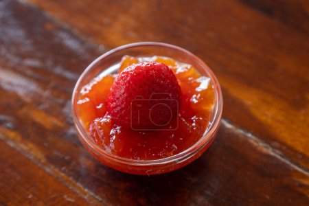 Foto de Un primer plano horizontal de una mermelada de fresa en un plato vidrioso con una fresa en ella sobre una mesa de madera - Imagen libre de derechos