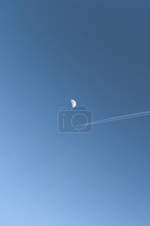 Photo for A vertical shot of a plane and its smoke trail seen in the sky with a half-moon in the back - Royalty Free Image