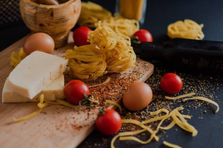 Photo for A view of pasta on a wooden board sprinkled with spice, tomatoes, cheese, two eggs and a grinding bowl - Royalty Free Image