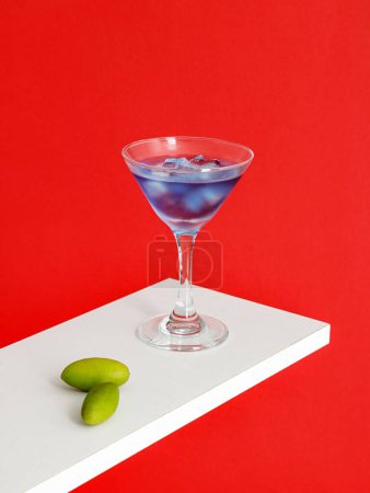 Téléchargez les photos : Un verre à cocktail Blue Moon et des olives sur surface blanche isolés sur fond rouge - en image libre de droit