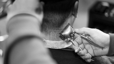 Foto de Un primer plano de las manos de un peluquero con una cortadora de pelo haciendo un corte de pelo para el cliente masculino, escala de grises - Imagen libre de derechos