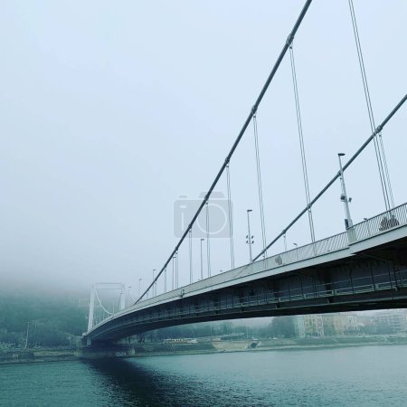 Téléchargez les photos : Vue panoramique à faible angle d'un pont au-dessus d'une rivière tranquille par une journée brumeuse - en image libre de droit