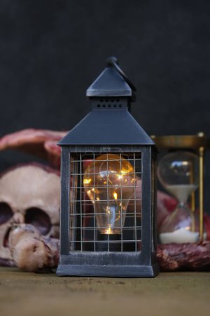 Foto de Una bombilla en una linterna negra, con un cráneo artificial y un reloj de arena en el fondo, sobre una mesa de madera, el concepto de decoración - Imagen libre de derechos