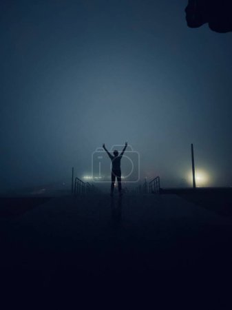 Foto de Un disparo vertical de una silueta de una persona alegre que muestra signos de paz en una noche de niebla - Imagen libre de derechos