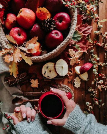Photo for A vertical top view of a woman holding a cup of coffee with a basket full of apples on a wooden table - Royalty Free Image