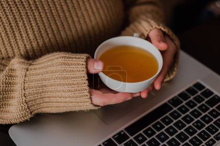 Foto de Un primer plano de una chica en un suéter acogedor sosteniendo una taza de té de limón sobre su computadora portátil en un café - Imagen libre de derechos