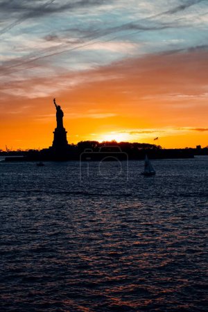 Photo for A vertical shot of sailboat in Hudson River in New York at Sunset and statue of Liberty silhouette in the background - Royalty Free Image