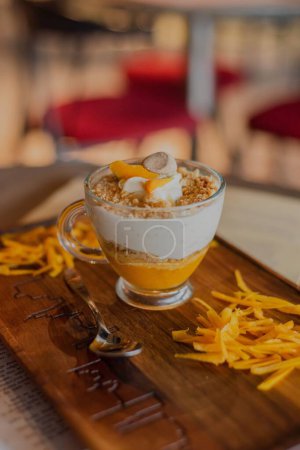 Photo for A vertical closeup of a glass of dessert with mango yogurt on a wooden board with mango slices, blurred background - Royalty Free Image