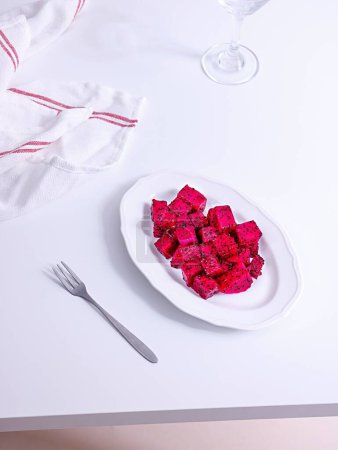 Photo for A vertical shot of pink dragon fruit cubes in a white plate with a fork and towel on a table - Royalty Free Image