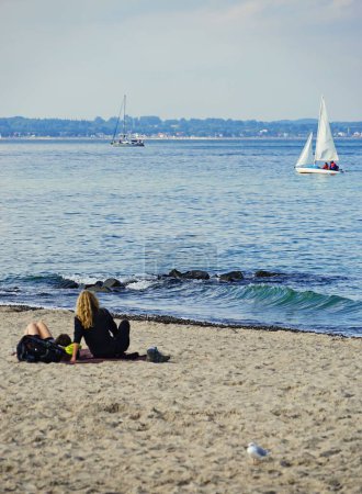 Foto de Una vista vertical de las personas que descansan en la playa de arena Travemuende con veleros en el fondo - Imagen libre de derechos