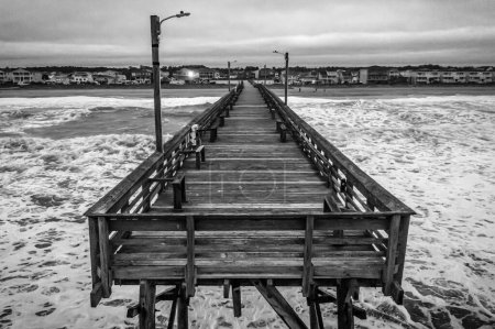 Photo for A high angle shot of a wooden pier on sea waves crashing against the beach shot in grayscale - Royalty Free Image