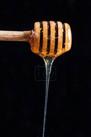 Photo for A vertical shot of honey dripping from a wooden honey dipper on a black background - Royalty Free Image