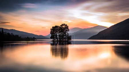 Foto de Una puesta de sol escénica sobre un lago con el reflejo de árboles y colinas en su superficie - Imagen libre de derechos