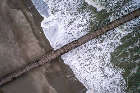 Foto de Una vista aérea de un muelle de madera sobre las olas del mar estrellándose contra la playa - Imagen libre de derechos