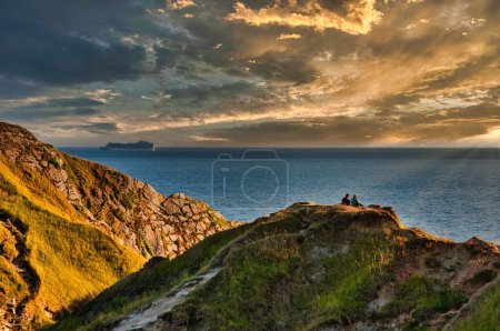 Photo for Dramatic cloudy sky over Lulworth Cove with a view of two people watching a peaceful waterscape from the rocky shore, UK - Royalty Free Image