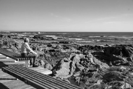 Téléchargez les photos : Une échelle de gris d'un vieil homme assis sur un banc sur une plage rocheuse, regardant les vagues de la mer - en image libre de droit