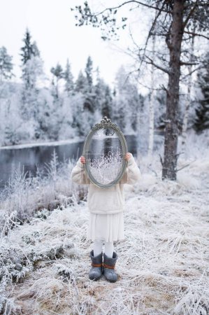 Photo for A vertical of a girl holding a mirror against her face with the cold winter forest in the background - Royalty Free Image