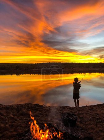 Photo for A vertical shot of a kid fishing near firewood from the shore with bright sunset sky in background - Royalty Free Image