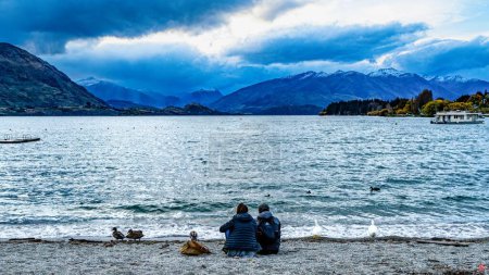 Téléchargez les photos : Un couple assis au bord du lac pittoresque Wanaka contre l'horizon des montagnes, Nouvelle-Zélande - en image libre de droit