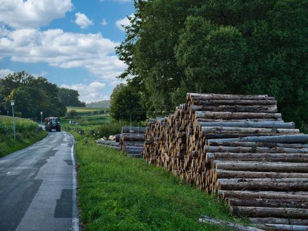 Téléchargez les photos : Une pile de bûches à côté d'une route avec un tracteur approchant en arrière-plan par une belle journée - en image libre de droit