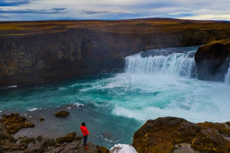 Téléchargez les photos : Un randonneur debout sur une falaise près de la cascade des Godafoos sous un ciel nuageux en Islande - en image libre de droit