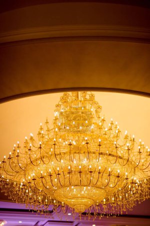 Photo for A low-angle shot of a luxurious chandelier hanging on a ceiling, with many lightbulbs turned on, decorated with hanging glassy ornaments - Royalty Free Image