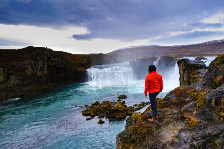 Téléchargez les photos : Un randonneur debout sur une falaise près de la cascade des Godafoos sous un ciel nuageux en Islande - en image libre de droit