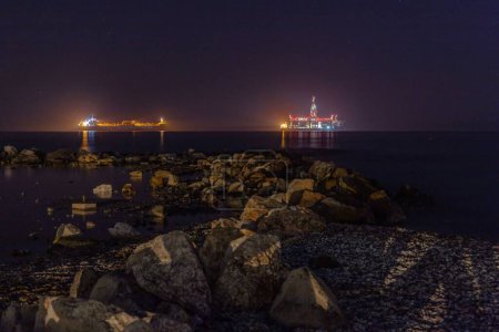 Foto de Una plataforma petrolífera en alta mar por la noche con luces reflejadas en el agua - Imagen libre de derechos