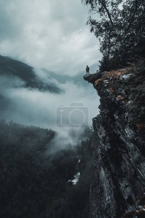 Photo for A vertical drone shot of a man on the edge of a rocky cliff facing the foggy forests - Royalty Free Image