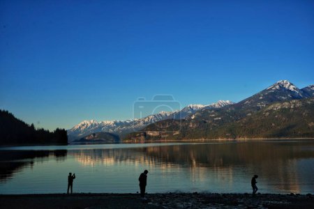 Foto de Las siluetas de los hombres en la costa de un lago rodeado de altas colinas bajo el cielo azul - Imagen libre de derechos