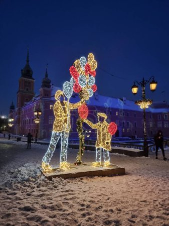 Téléchargez les photos : Travaux légers faits de lampes colorées sur la place principale enneigée de la vieille ville de Varsovie dans la soirée. iI montre un homme avec des ballons et un enfant. - en image libre de droit