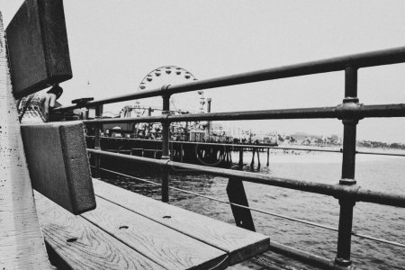 Téléchargez les photos : Un cliché en échelle de gris d'un banc en bois sur la jetée du port de Santa Monica, en Californie, avec la mer et la grande roue en face à Los Angeles, États-Unis - en image libre de droit