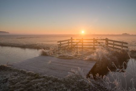 Foto de Un río helado a lo largo de los campos cubiertos de nieve, y un puente de madera congelado durante el amanecer en invierno - Imagen libre de derechos