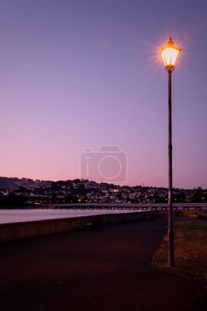 Téléchargez les photos : Une lanterne de rue illuminée brille à côté de la rivière Teign contre le ciel violet à Shaldon Devon - en image libre de droit