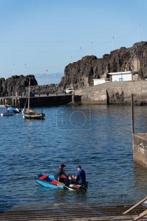 Téléchargez les photos : Deux hommes dans un petit bateau, ramant à Madère, Portugal, avec des bateaux dans la jetée rocheuse en arrière-plan, par une journée ensoleillée - en image libre de droit