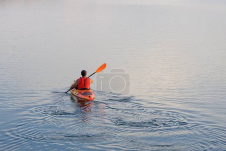 Téléchargez les photos : Un homme sur un kayak pagayant dans un lac calme sous un éclairage doux au coucher du soleil - en image libre de droit