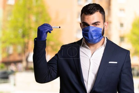 Téléchargez les photos : Un bel homme d'affaires avec un masque chirurgical et des gants tenant un tube à sang dans un parc - en image libre de droit