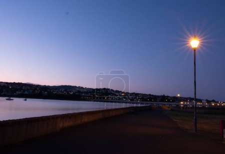 Téléchargez les photos : Une lanterne de rue illuminée brille à côté de la rivière Teign contre le ciel violet à Shaldon Devon - en image libre de droit