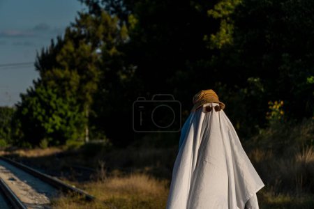 Foto de Una persona cubierta por una sábana blanca, con gafas de sol y sombrero, en apariencia de fantasma, en un jardín durante el día en Guadalajara, México - Imagen libre de derechos
