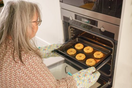 Photo for Older white-haired woman putting a tray of cookies in the oven - Royalty Free Image